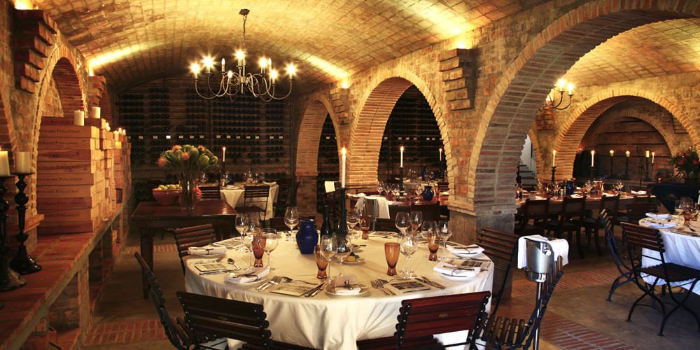 Rustikaler dezent beleuchteter Gewölbe-Weinkeller mit gedeckten Holztischen und besonderem Ambiente