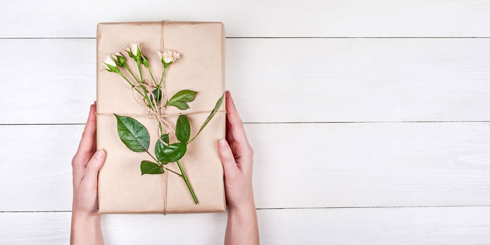 Ein schön verpacktes Geschenk verziert mit einem Rosenstiel wird von zwei Händen vor einer Holzplatte gehalten