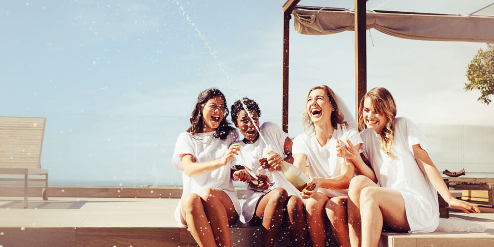 Vier fröhliche Frauen sitzen auf einer Terrasse und entkorken eine Flasche Sekt