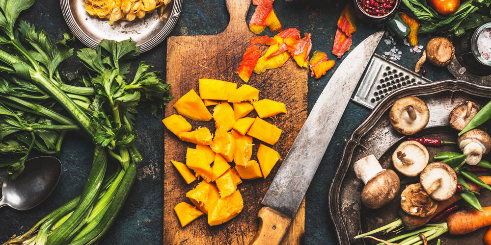 Kürbis liegt geschnitten auf einem Holzbrett neben Pilzen, Mohrrüben, Staudensellerie und Gewürzen auf einem Tisch